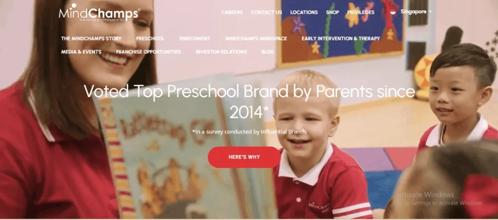 A smiling preschool teacher reading a book to two young students wearing red polo uniforms in a classroom, featured on the MindChamps website banner.