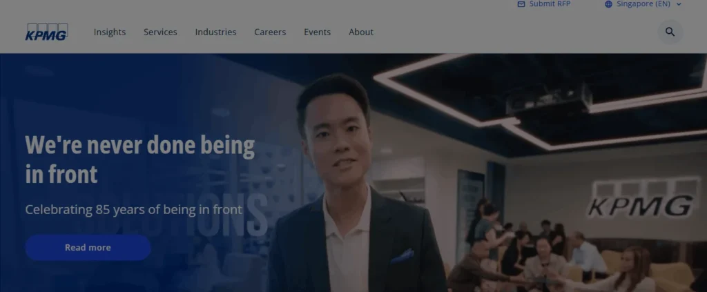 Screenshot of the KPMG Singapore website homepage. The website header features the KPMG logo alongside navigation links like Insights, Services, Industries, Careers, Events, and About. The main banner shows a professional man in a suit standing in front of a modern office environment with a large KPMG logo on the wall and a group of people sitting in the background. The text overlay reads, "We're never done being in front" and "Celebrating 85 years of being in front," with a blue "Read more" button below.