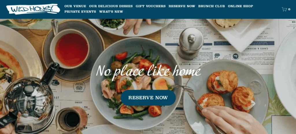 A website header for Wild Honey restaurant featuring a dark blue navigation bar and a prominent banner image showing an overhead view of a brunch table setting. White cursive text overlays the image reading "No place like home" above a blue "RESERVE NOW" button.