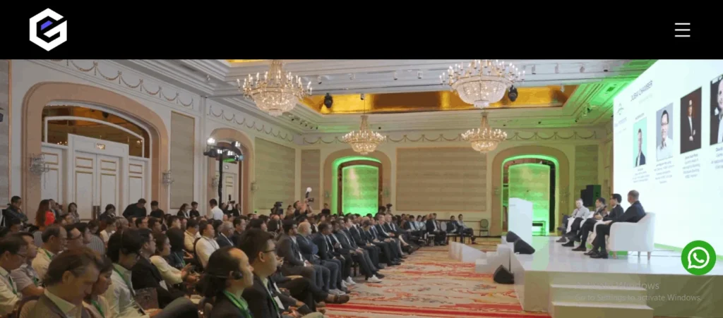 Wide shot of a corporate conference in a grand, classically designed ballroom with crystal chandeliers. A panel of speakers sits on a raised stage next to a large presentation screen, addressing a seated audience.
