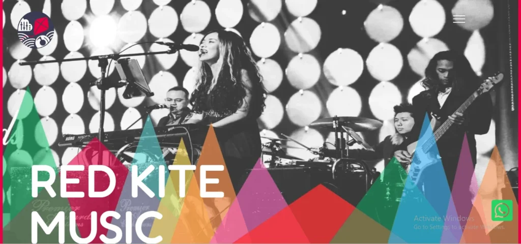 A website homepage banner for Red Kite Music featuring a black-and-white photo of a live band performing on stage. The band includes a female lead vocalist singing, a keyboardist, a drummer, and a bass guitarist, set against a circular bokeh background. Bold, colorful geometric triangles in red, orange, green, and purple run along the bottom with the text "RED KITE MUSIC" overlaid.