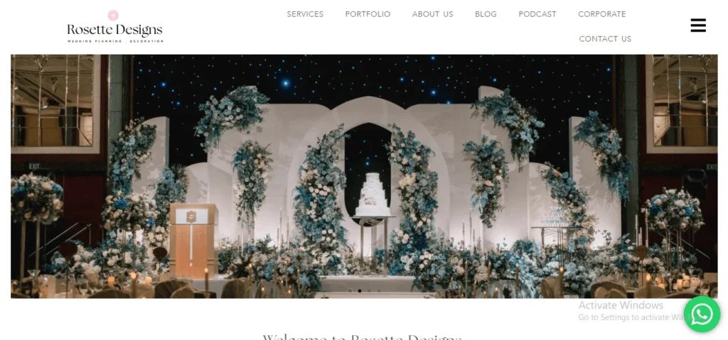 Website banner for Rosette Designs showing an elegant wedding stage setup with a white tiered wedding cake, a gold podium, and elaborate blue and white floral decorations against a starry night backdrop.
