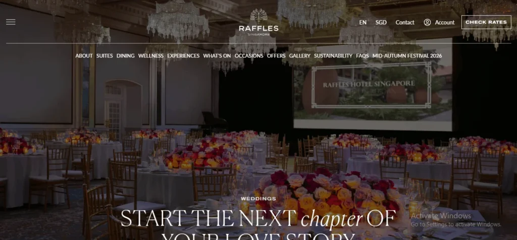 A grand ballroom at Raffles Hotel Singapore set up for a wedding banquet, featuring elegant round tables adorned with large red, pink, and yellow floral centerpieces, crystal chandeliers overhead, and a large projection screen in the background.